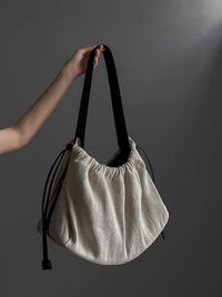 Beige handbag with black strap held by a hand against a gray background