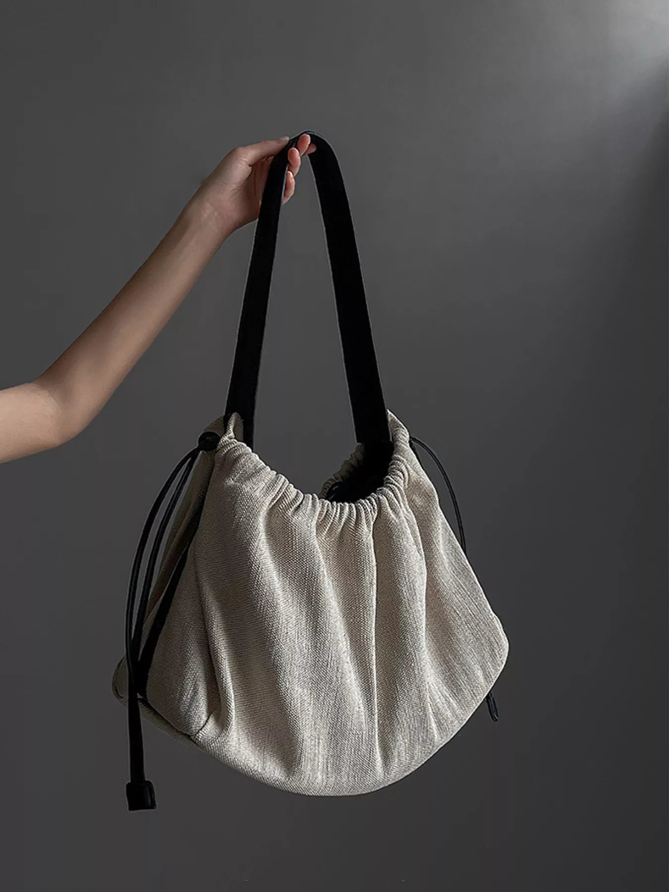 Beige handbag with black strap held by a hand against a gray background