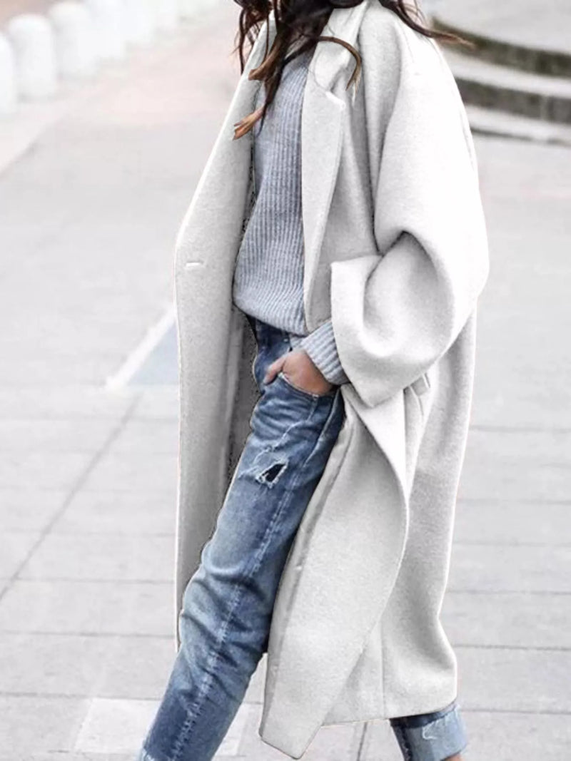Person wearing a long gray coat over a light blue sweater and jeans on a blurred background