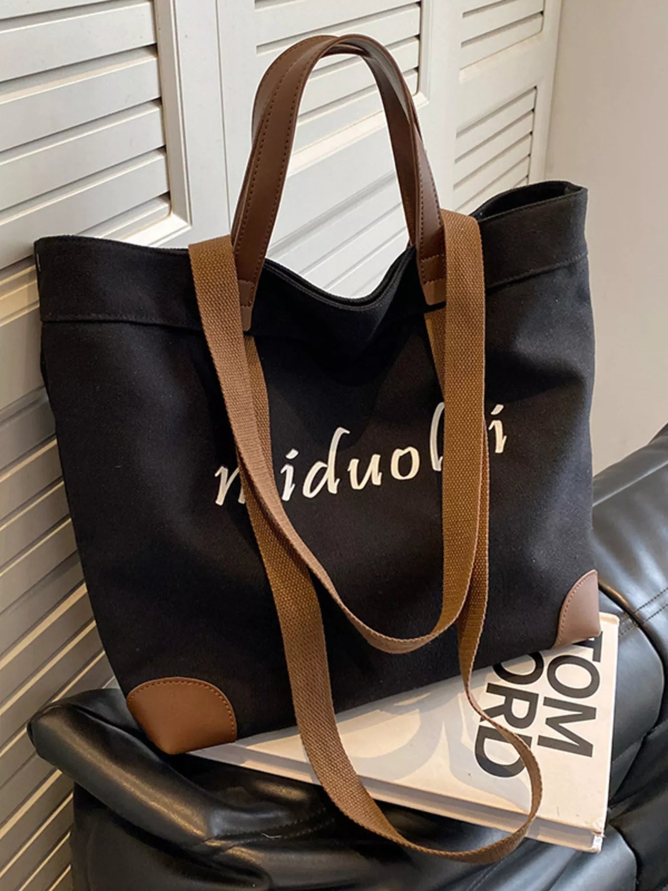 Black tote bag with brown handles on a chair next to a book