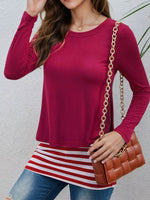 A woman wearing a long-sleeve crewneck top in a deep magenta pink, styled with a red and white striped tunic, blue jeans, and a brown quilted crossbody bag with a gold chain strap.