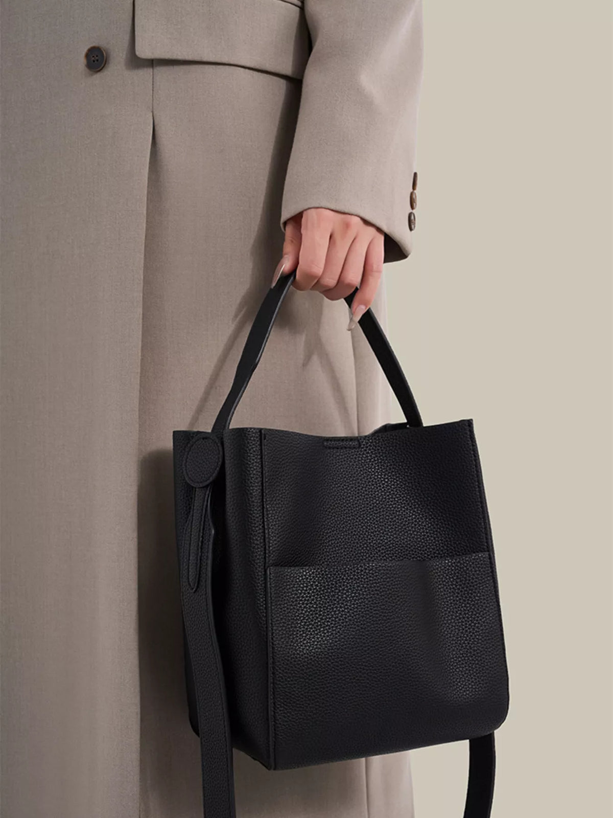 A person in a light beige trench coat holds a black pebbled leather bucket bag with top handles and a long shoulder strap against a neutral beige background.