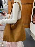 A person wearing a white ribbed turtleneck and light pink pants carries a large slouchy tan suede tote bag over their shoulder in an outdoor urban setting.
