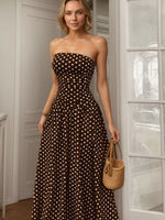 Woman wearing a brown strapless dress with white polka dots, holding a woven handbag indoors.