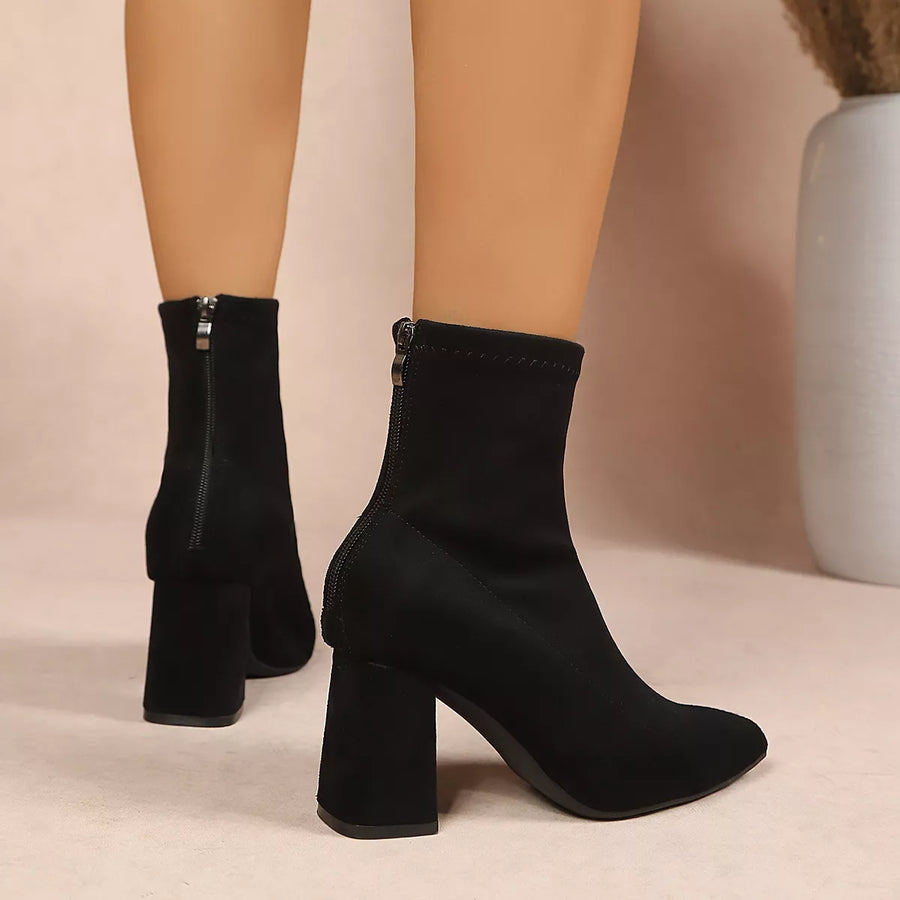 Black ankle boots with a block heel and side zipper, shown on a model's feet against a neutral beige background.