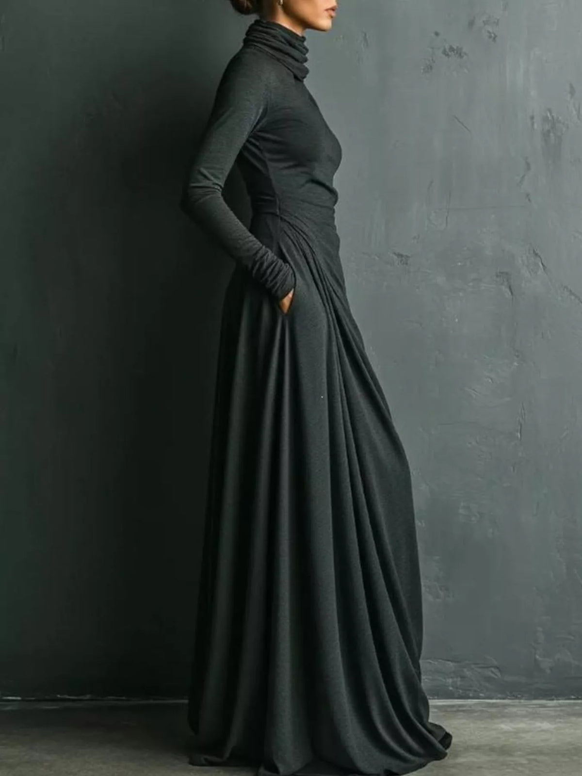 A woman wearing a long-sleeve, high-neck, floor-length black dress with gathered draping at the waist, standing against a dark textured wall.