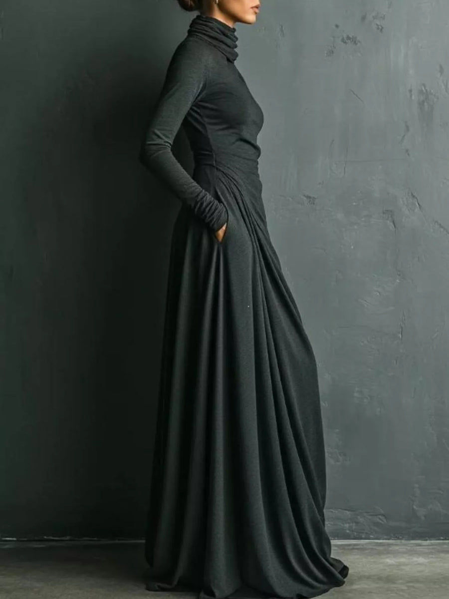 A woman wearing a long-sleeve, high-neck, floor-length black dress with gathered draping at the waist, standing against a dark textured wall.