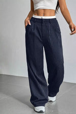 A person wearing loose navy pinstripe pants with a white waistband, paired with a white crop top and white sneakers, standing against a light gray wall.