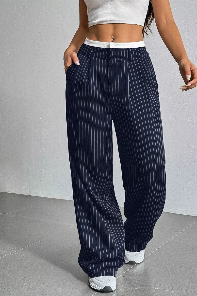 A person wearing loose navy pinstripe pants with a white waistband, paired with a white crop top and white sneakers, standing against a light gray wall.
