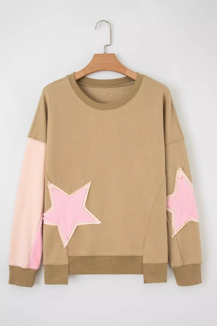 A beige crewneck sweatshirt hanging on a wooden hanger against a light gray wall. The sweatshirt features light pink star-shaped patches on the front and sleeves, with light pink elbow patches and ribbed beige cuffs and hem.