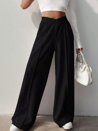 Person wearing black wide-leg pants and a white top, holding a white handbag against a plain background.