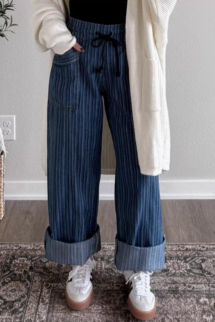 Person wearing blue striped pants and white sneakers in a room with a gray wall and patterned rug.