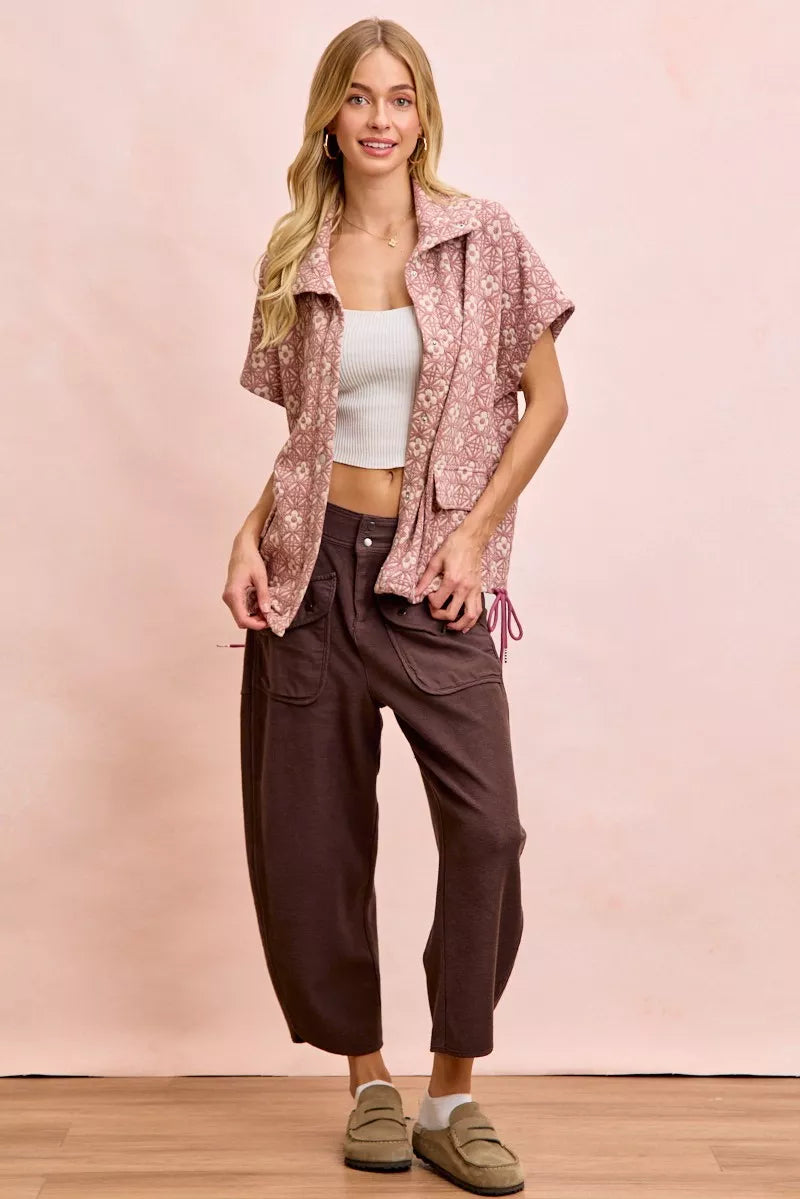 Woman wearing a floral shirt, white top, and brown pants against a pink background