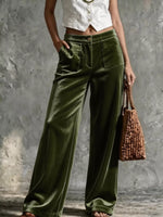 A model wearing high-waisted, wide-leg olive green velvet pants, styled with a white cropped vest, holding a brown woven handbag against a textured gray concrete wall.