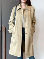 A woman wearing a mid-length beige trench coat over a light top and blue jeans, standing indoors against a plain light wall.