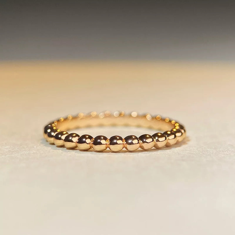 A delicate gold beaded ring resting on a light beige textured surface with a soft gray background.