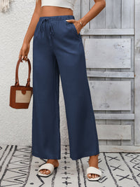 Person wearing blue wide-leg pants with a white top and sandals, holding a brown handbag on a patterned rug.