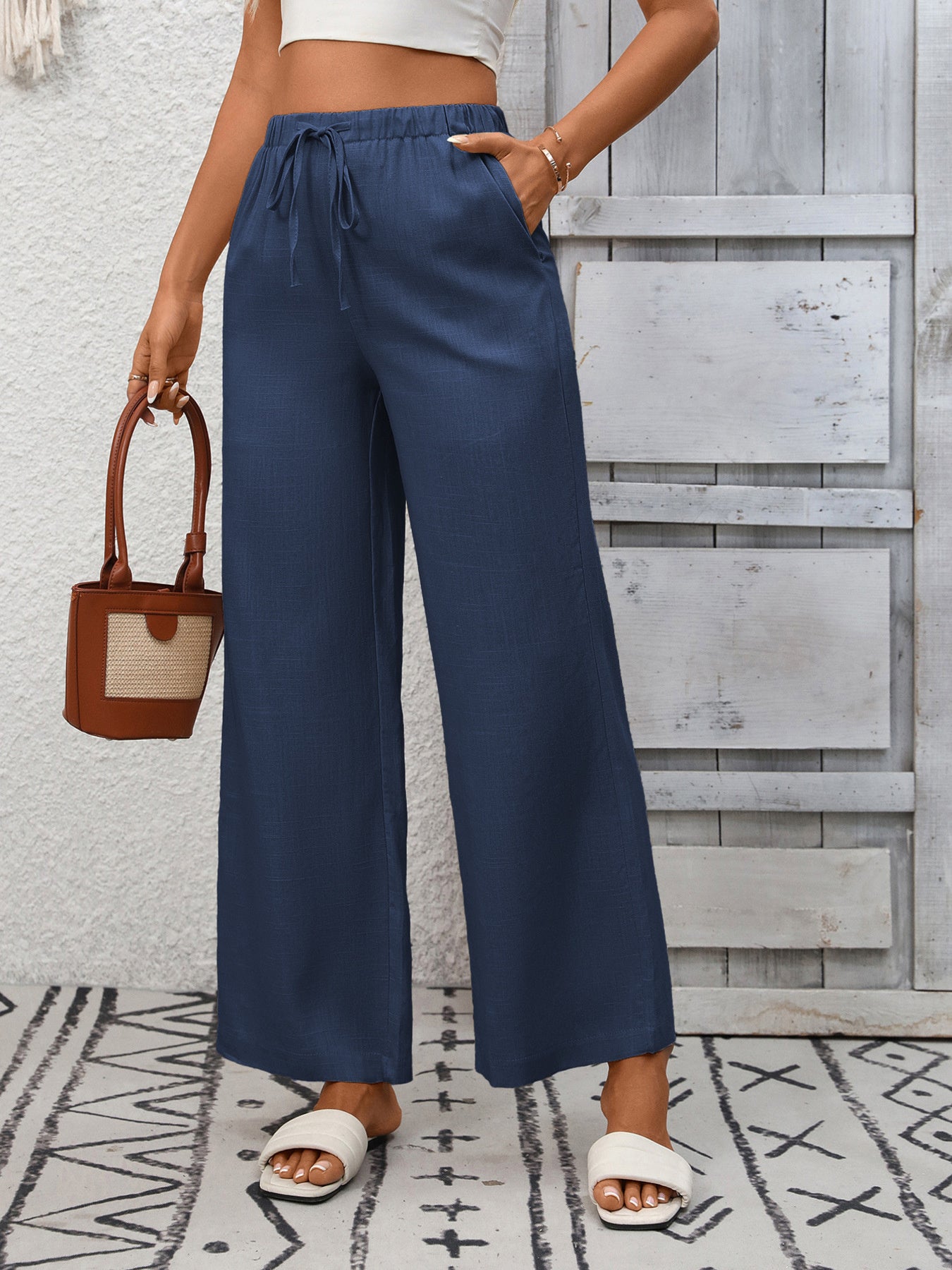 Person wearing blue wide-leg pants with a white top and sandals, holding a brown handbag on a patterned rug.
