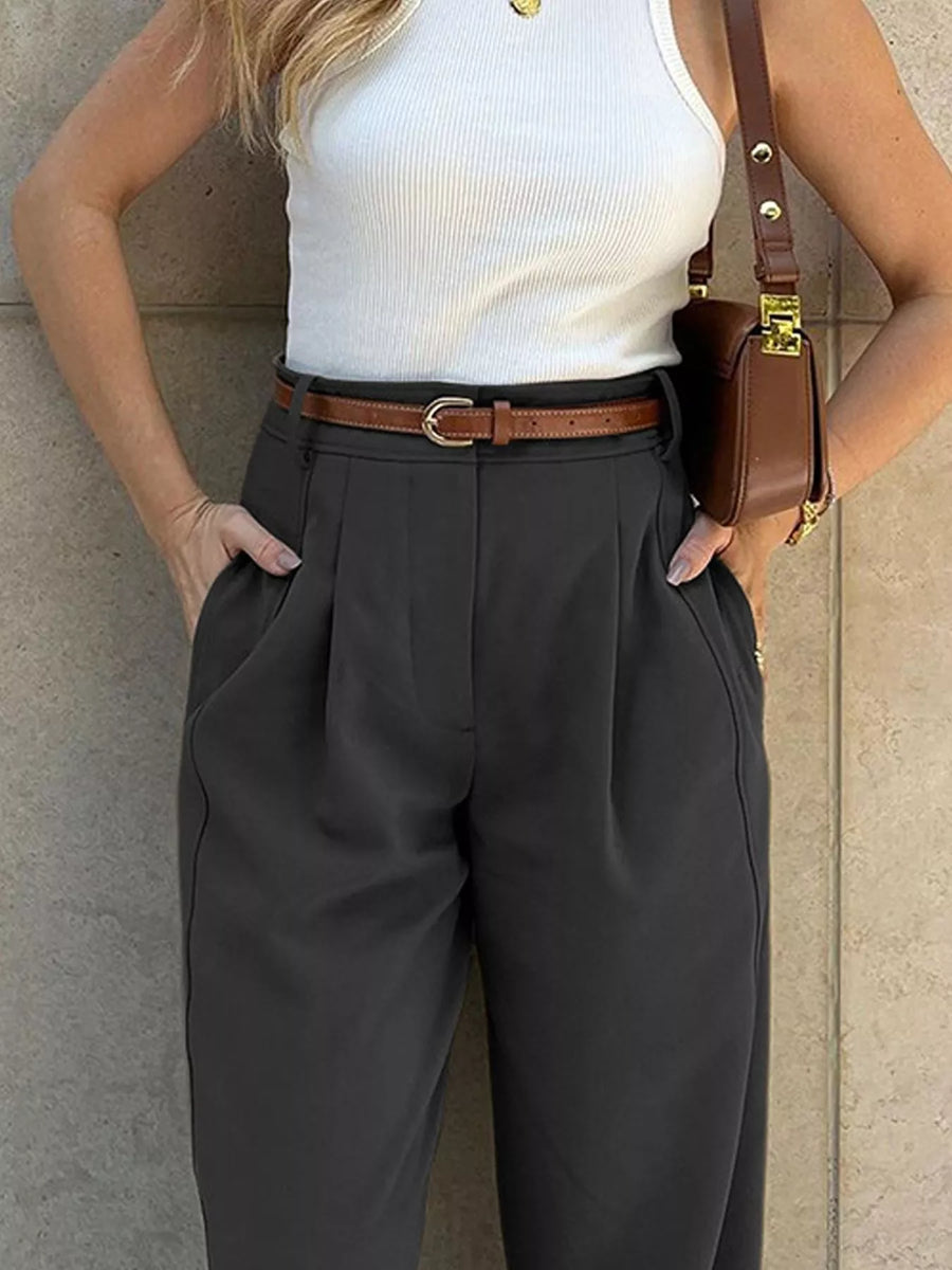 A woman wearing dark charcoal gray pleated trousers with a brown leather belt, paired with a white ribbed tank top and a brown crossbody bag, standing against a beige stone wall.