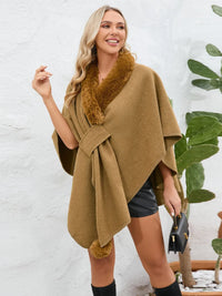 Woman wearing a brown poncho with fur trim against a white wall.