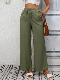 Person wearing green wide-leg pants with a white top and sandals, standing against a wooden panel wall.