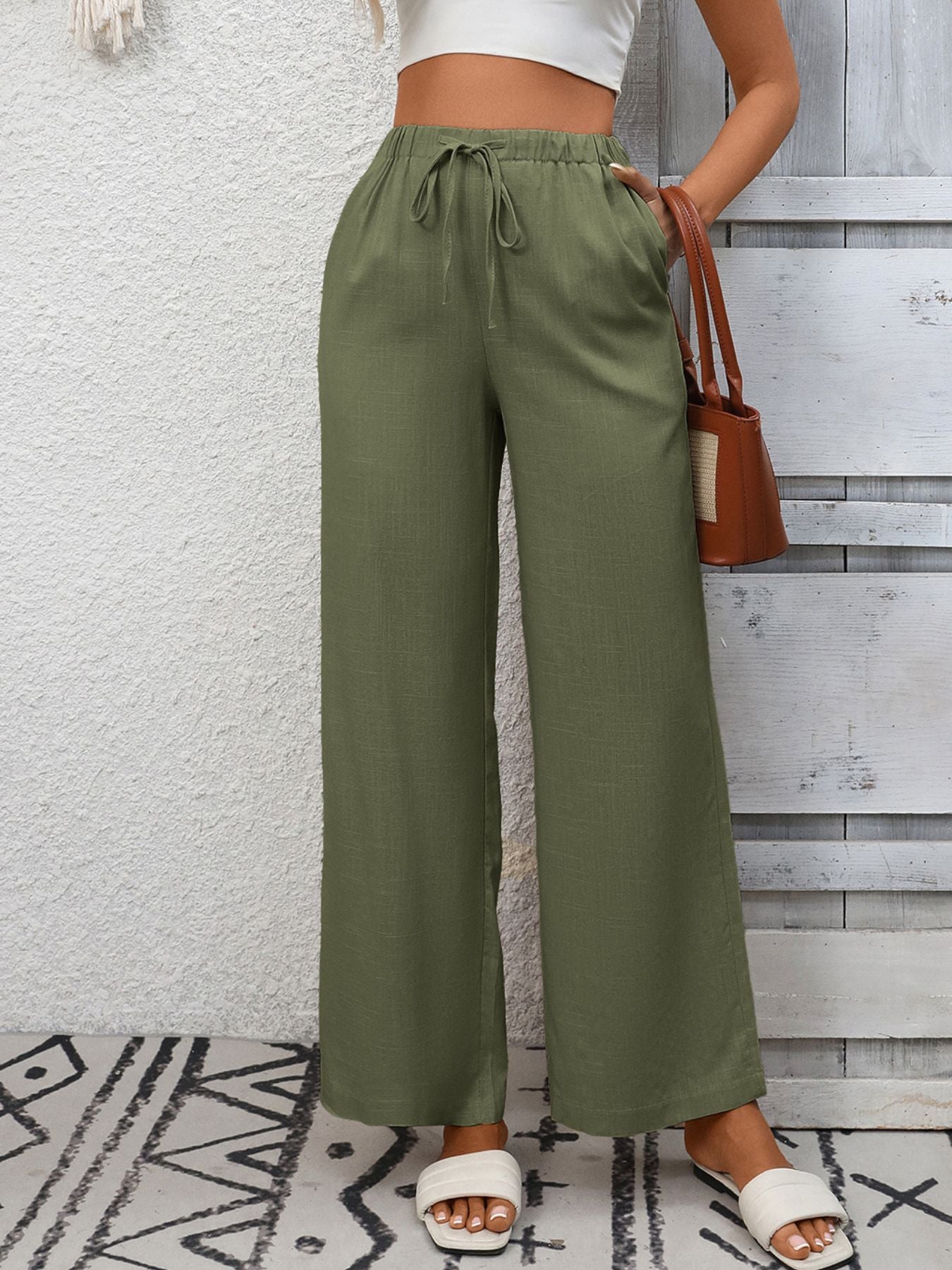 Person wearing green wide-leg pants with a white top and sandals, standing against a wooden panel wall.