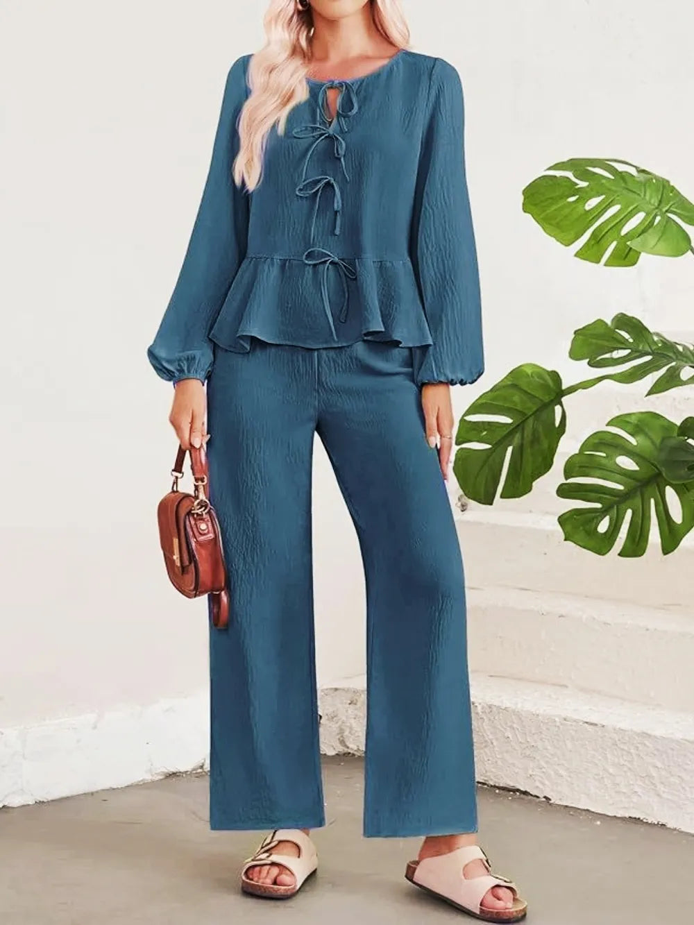 Woman wearing a blue outfit with a white wall and green leaves in the background