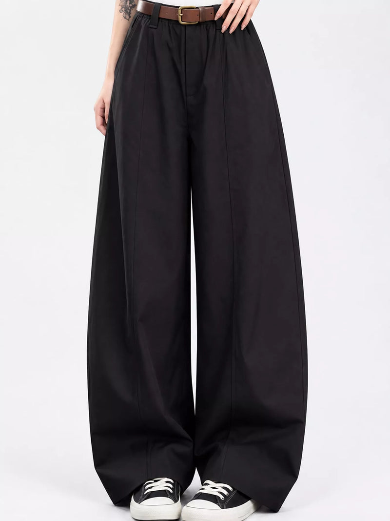 Black wide-leg trousers with a high waist, styled with a brown belt, black and white sneakers, and a visible model's hands and lower torso against a plain white studio background.