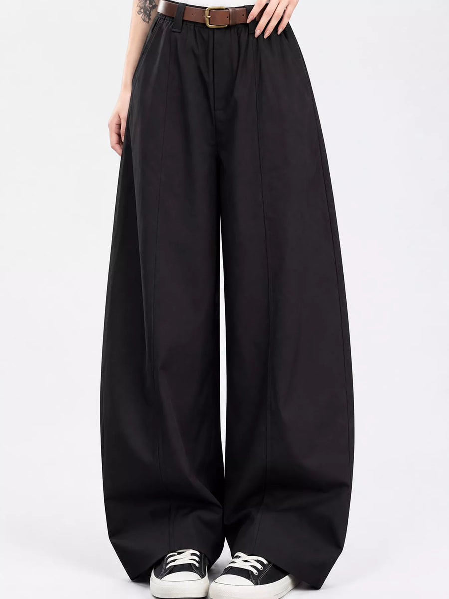 Black wide-leg trousers with a high waist, styled with a brown belt, black and white sneakers, and a visible model's hands and lower torso against a plain white studio background.