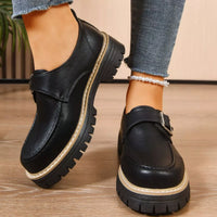 Black leather shoes with thick soles worn by a person on a wooden floor.