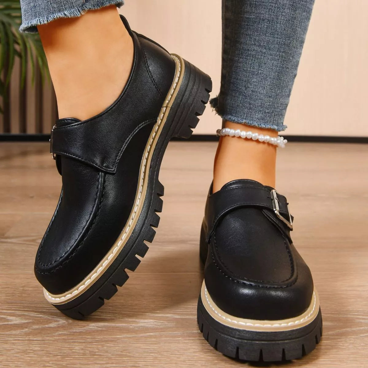 Black leather shoes with thick soles worn by a person on a wooden floor.