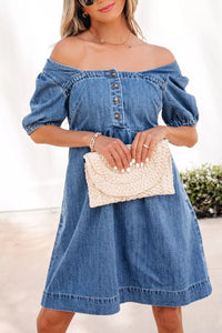 Woman wearing a blue denim dress holding a beige clutch against a white background