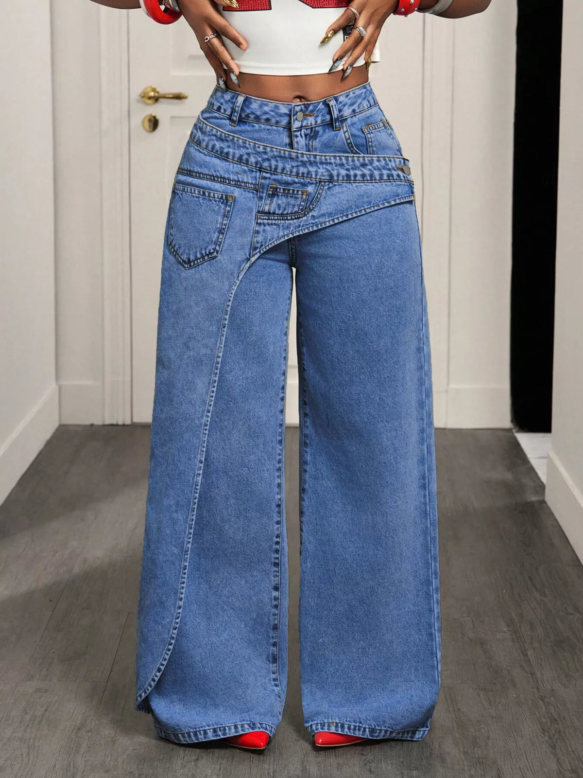 A person wearing high-waisted, wide-leg blue denim jeans with a diagonal seam detail down the front of one leg, paired with a white crop top and red shoes, standing in an indoor setting with a gray floor and white door.