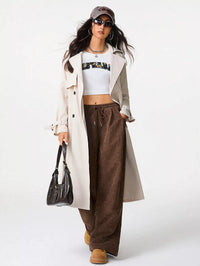 Woman wearing a long beige coat, white crop top, brown wide-leg pants, and tan boots on a white background