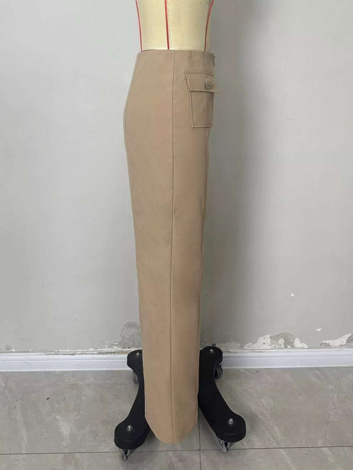A beige pencil skirt displayed on a dress form with black feet, set against a plain gray wall and tiled floor.