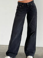A person wearing dark washed wide-leg jeans with white sneakers, shown from the waist down against a plain light gray wall and concrete floor.