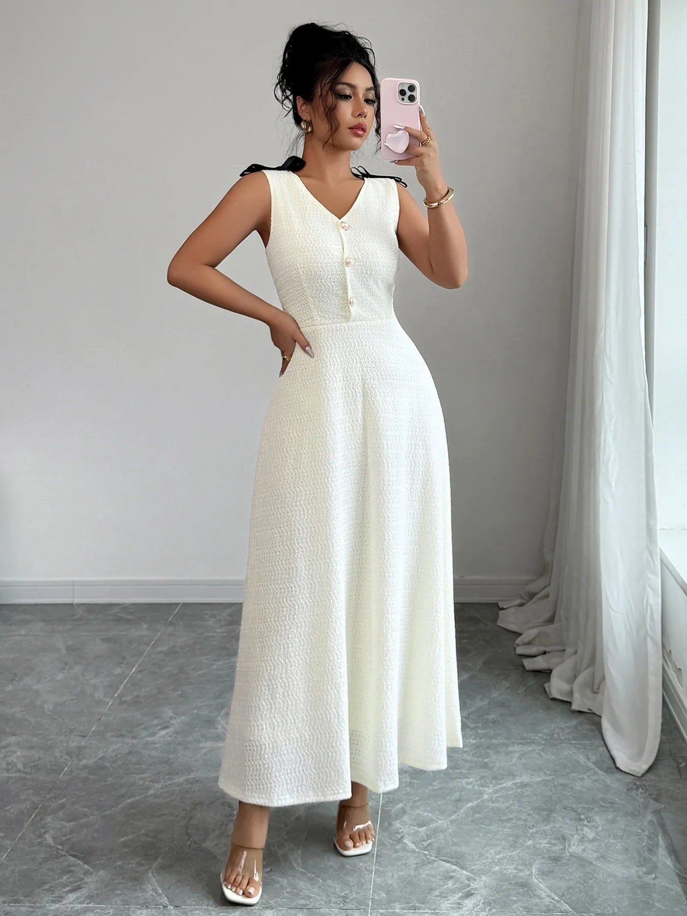 Woman in a white sleeveless dress taking a mirror selfie in a minimalistic room.