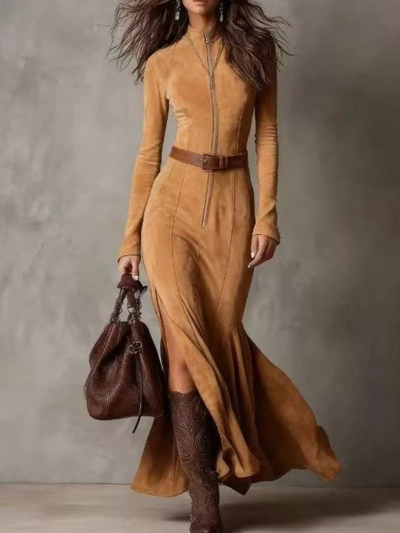 Woman wearing a long brown dress with a belt, holding a matching brown handbag against a neutral background.