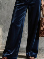 A close-up of a person wearing dark navy blue velvet wide-leg pants, holding a brown woven handbag against a textured gray concrete wall.