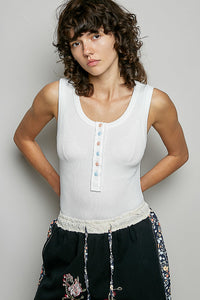 Woman wearing a white sleeveless top with buttons and black pants with floral embroidery against a plain background