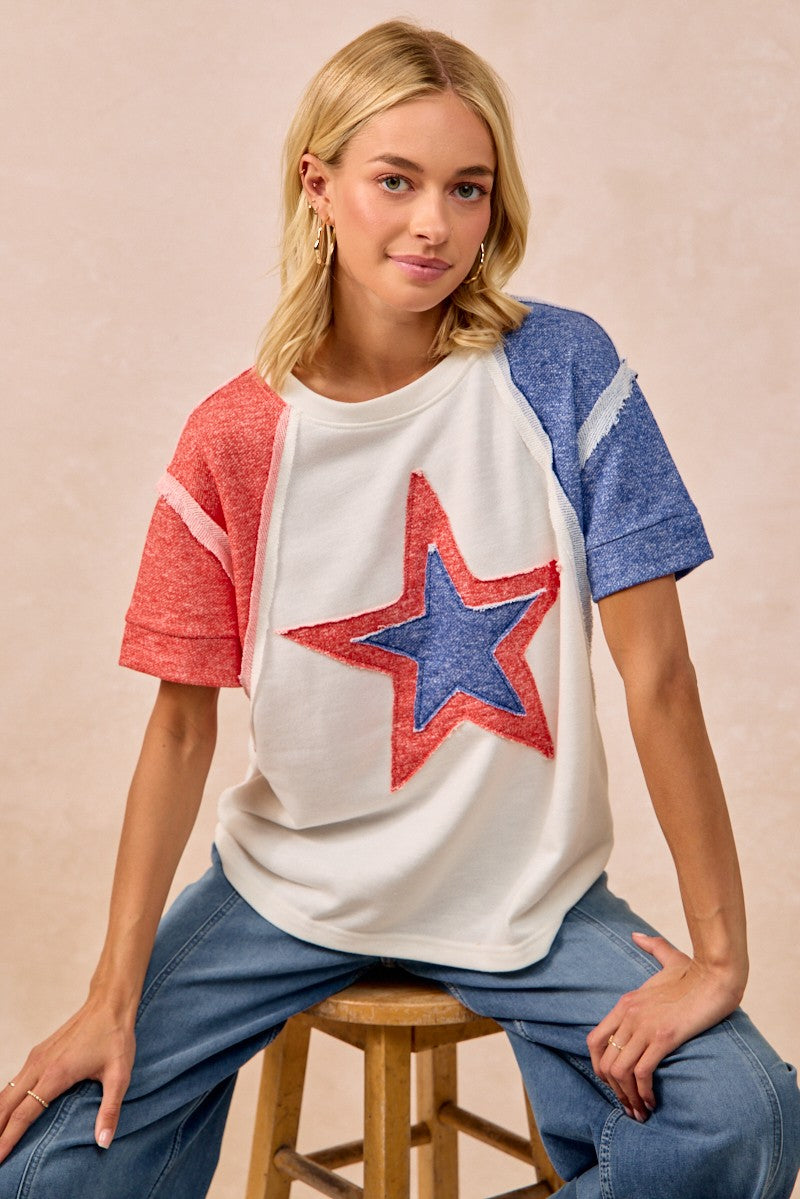 Woman wearing a colorful star-themed shirt sitting on a stool against a plain background