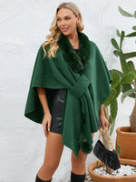 Woman wearing a green poncho with fur trim against a white wall.