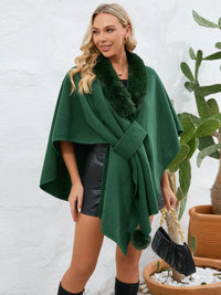 Woman wearing a green poncho with fur trim against a white wall.
