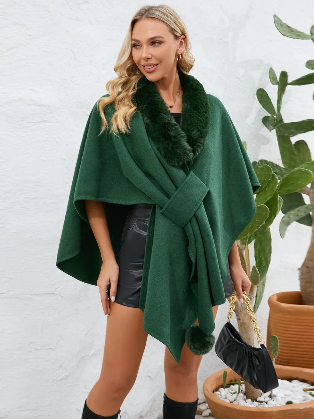 Woman wearing a green poncho with fur trim against a white wall.