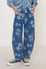 Person wearing blue jeans with white floral patterns and a denim jacket on a plain background