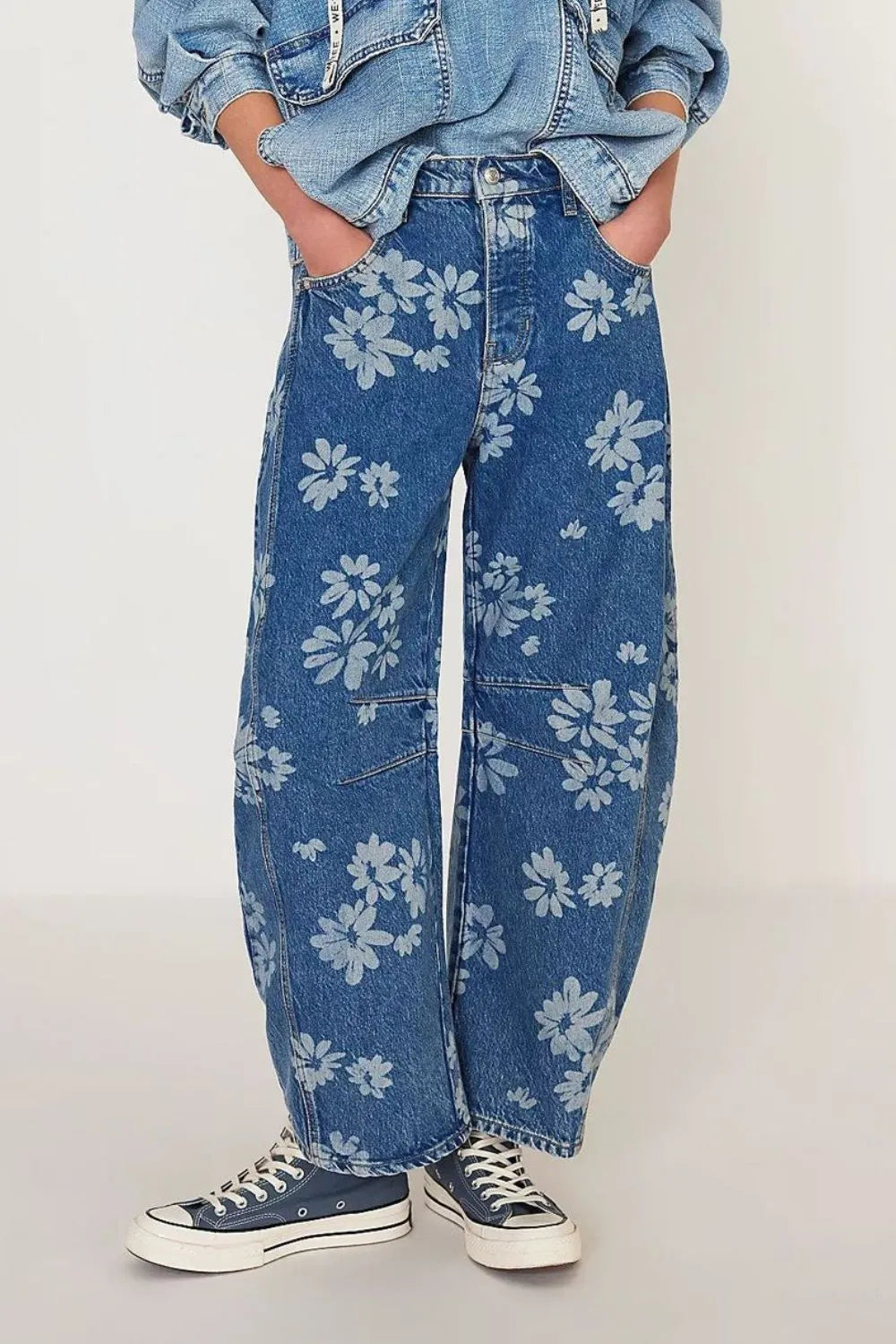 Person wearing blue jeans with white floral patterns and a denim jacket on a plain background