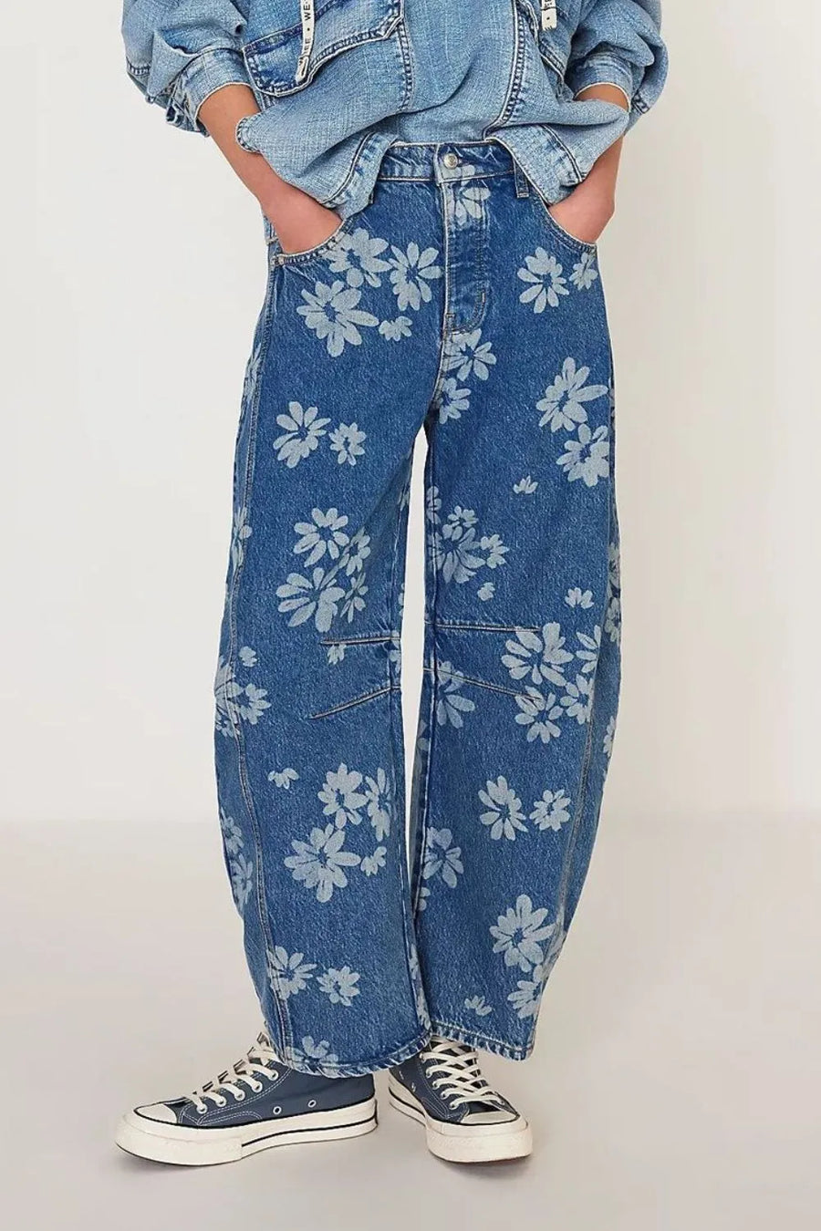 Person wearing blue jeans with white floral patterns and a denim jacket on a plain background