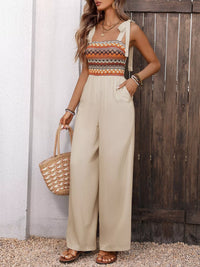 Woman wearing a sleeveless top with a geometric pattern and beige wide-leg pants, standing against a wooden fence.