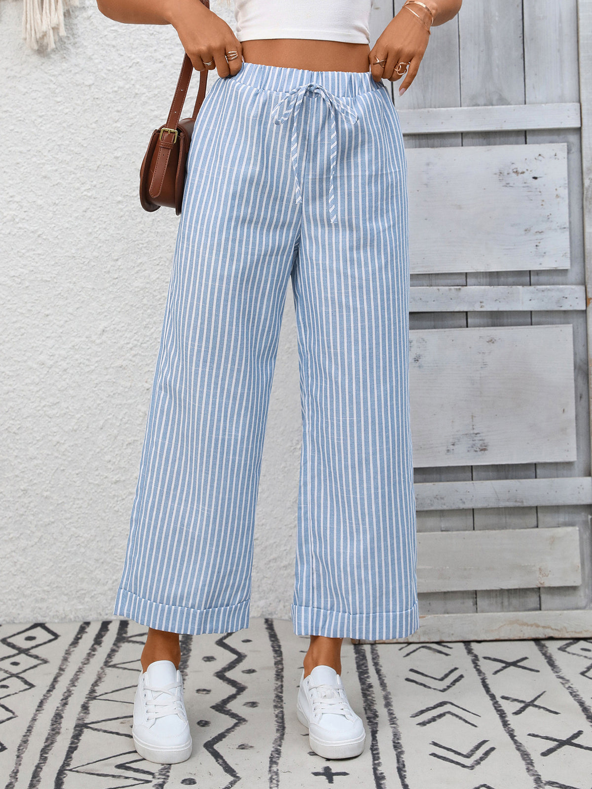 Person wearing blue and white striped pants with a white top and white sneakers, standing against a wooden wall.
