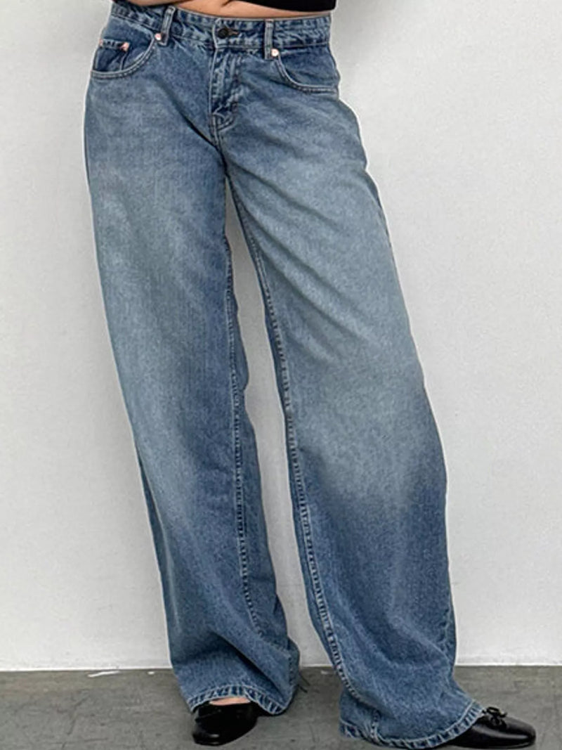 A person wearing light blue wide-leg denim jeans with black shoes, standing against a plain light gray wall.